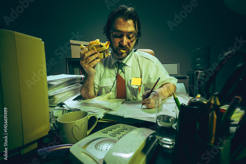 Obraz na płótnie Exhausted office worker eats sandwich, writing at messy, dimly lit desk, showing extreme burnout and inability to take proper break. Concept of overwork, no work-life balance, and stress. w salonie