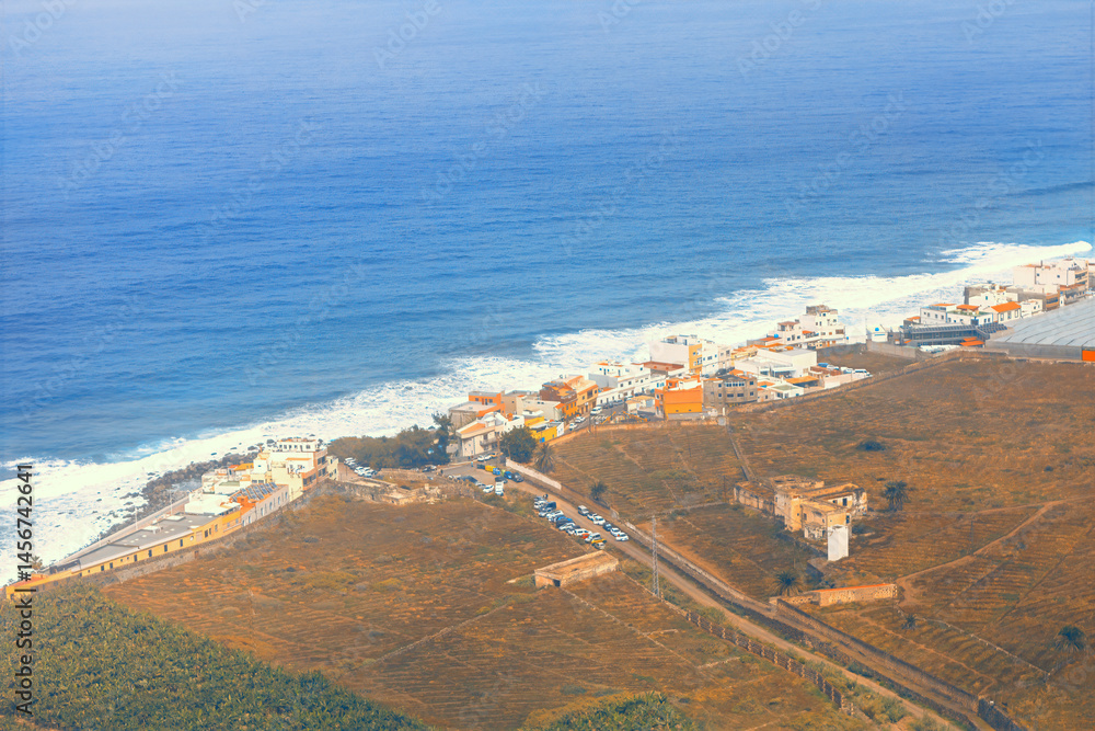 View of coastal area on Gran Canaria, featuring a small village with colorful buildings nestled along a rugged shoreline. Ocean waves crash against the coast and road extending inland from settlement