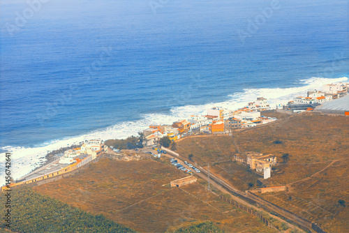 View of coastal area on Gran Canaria, featuring a small village with colorful buildings nestled along a rugged shoreline. Ocean waves crash against the coast and road extending inland from settlement