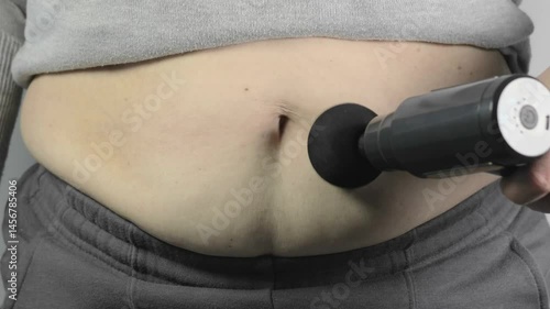 Woman doing anti cellulite massage on her fat belly with percussion massage gun. Overweight problem on stomach. Physiotherapy, self-massage tool. Healthcare. Close up, 4k footage.