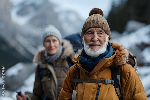 Two hikers enjoy their winter adventure in the mountains. The elderly man stands in the foreground, wearing a warm jacket and hat, while the woman, slightly behind, looks happily at the camera