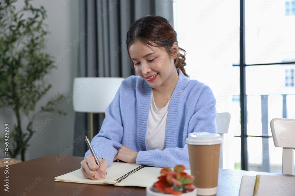 Focused Asian young woman sitting at desk, writing in notebook with pen. She wears cozy blue sweater, appearing thoughtful and concentrated. Bright and calm, ideal for studying, journaling, or plannin Focused Asian young woman sitting at desk, writing in notebook with pen. She wears cozy blue sweater, appearing thoughtful and concentrated. Bright and calm, ideal for studying, journaling, or plannin