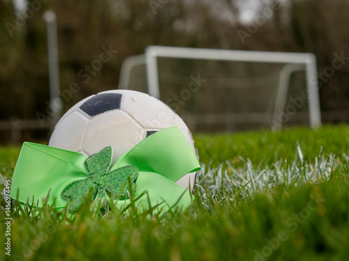Obraz na płótnie Green bow with Irish shamrock by a classic football or soccer ball on a green grass training field. Goal post in the background. Supporting national team of Ireland concept. w salonie