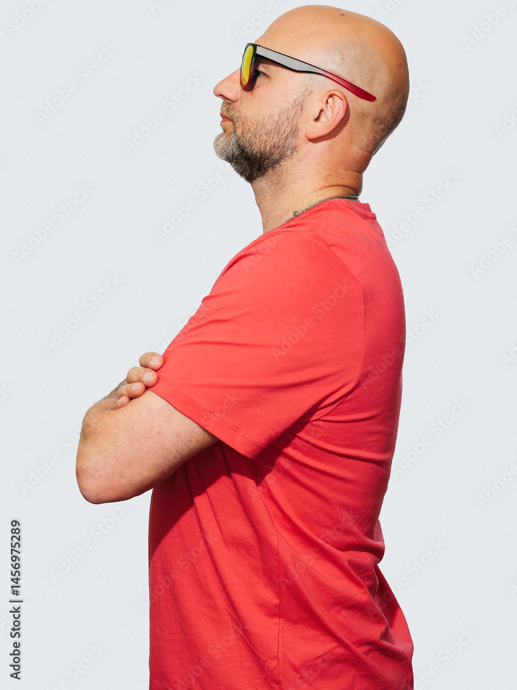 A man in a red shirt is standing with his arms crossed. He has a serious expression on his face, light color background. Male in his 40s with grey beard and bold head, slim body type.