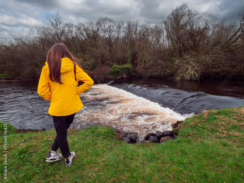 Female tourist in yellow jacket looking at a small river with waterfall. Travel and tourism. Enjoy nature scenery theme. Girl standing by a river with nice view.