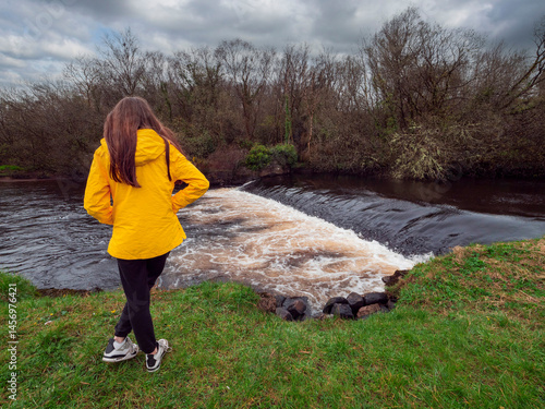 Obraz na płótnie Female tourist in yellow jacket looking at a small river with waterfall. Travel and tourism. Enjoy nature scenery theme. Girl standing by a river with nice view. w salonie