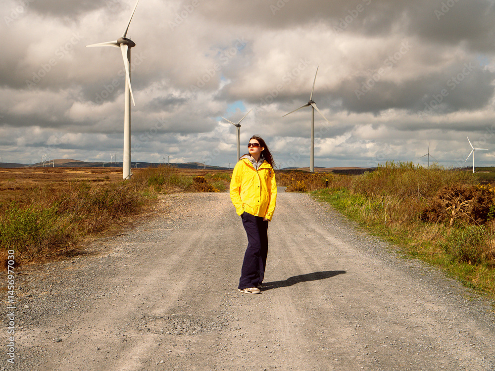 Young teenager girl in yellow jacket standing on a road, Wind turbines and vast wild fields in the background. Learning clean and green energy production. Warm sunny day.