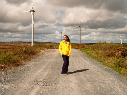 Obraz na płótnie Young teenager girl in yellow jacket standing on a road, Wind turbines and vast wild fields in the background. Learning clean and green energy production. Warm sunny day. w salonie