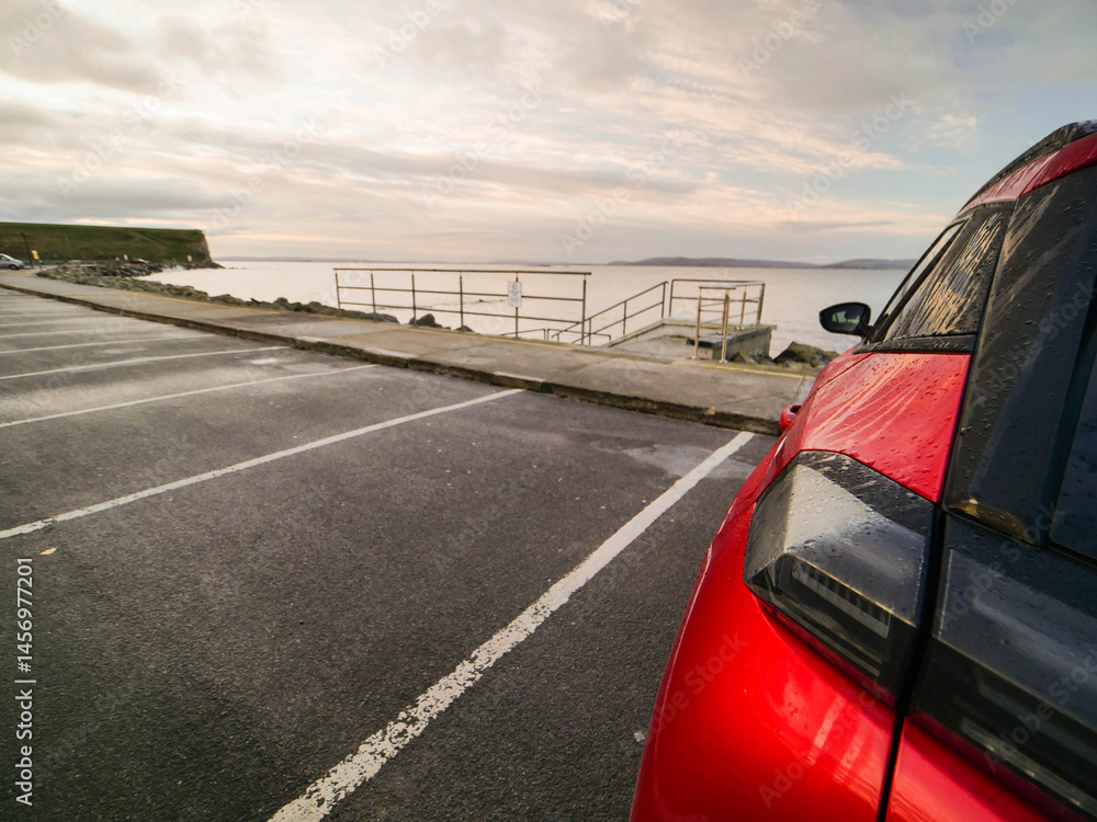 Red car parked in car park with stunning view on nature scenery with ocean and mountains and cloudy sky. Silverstrand area, Galway city, Ireland. Rent motor for a tourist trip concept.