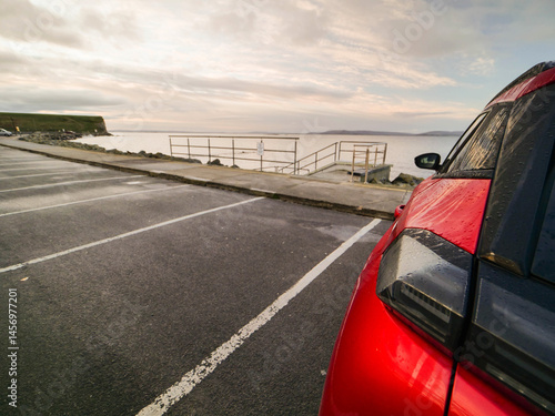 Obraz na płótnie Red car parked in car park with stunning view on nature scenery with ocean and mountains and cloudy sky. Silverstrand area, Galway city, Ireland. Rent motor for a tourist trip concept. w salonie