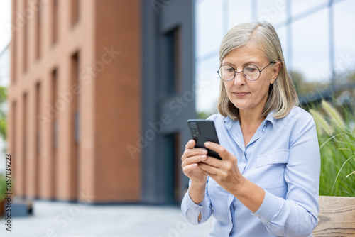 Obraz na płótnie A mature woman with glasses, sitting outdoors, focused on her smartphone. She is engaged in messaging or browsing on her mobile device. w salonie