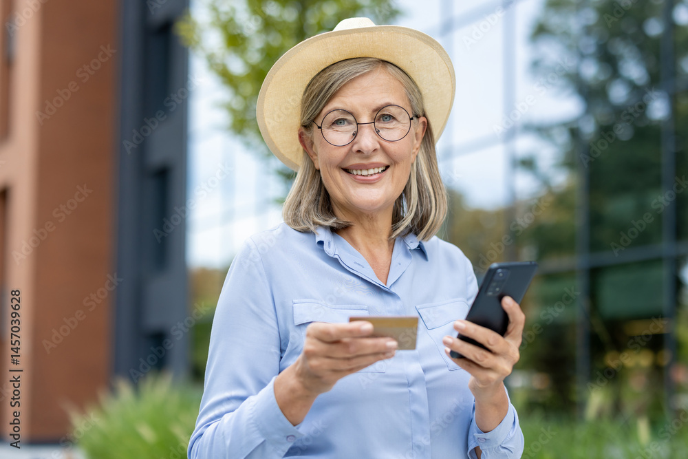 A mature woman smiles while holding a credit card and a smartphone outdoors, possibly shopping online. She wears a stylish hat.