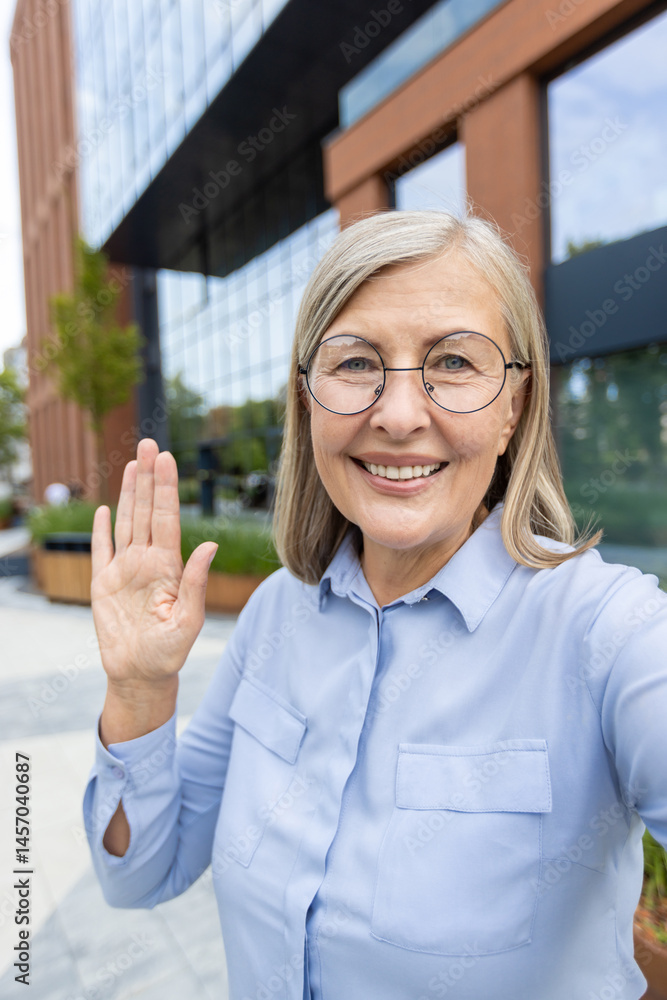 A friendly senior woman waves in front of a modern office building, smiling and welcoming.