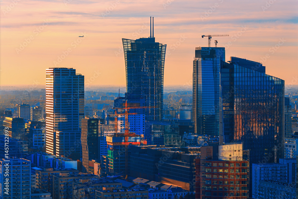 Dynamic sunset view of Warsaw modern skyline, with high-rise glass buildings and cranes symbolizing growth. A plane in the sky adds motion to this vibrant European business hub