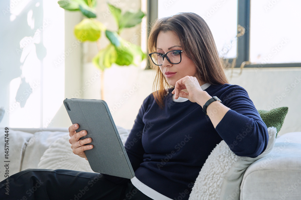 Serious middle aged woman looking at digital tablet sitting on sofa at home