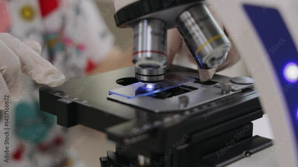Gloved hands carefully position a microscope slide on a microscope stage, illuminated from below Gloved hands carefully position a microscope slide on a microscope stage, illuminated from below