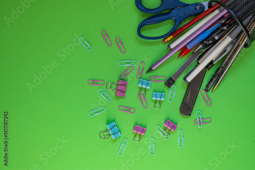 This image features an assortment of office supplies such as pens, pencils, scissors, binder clips, and paper clips spilling out of a mesh holder onto a vivid green surface. The mix of colors and item