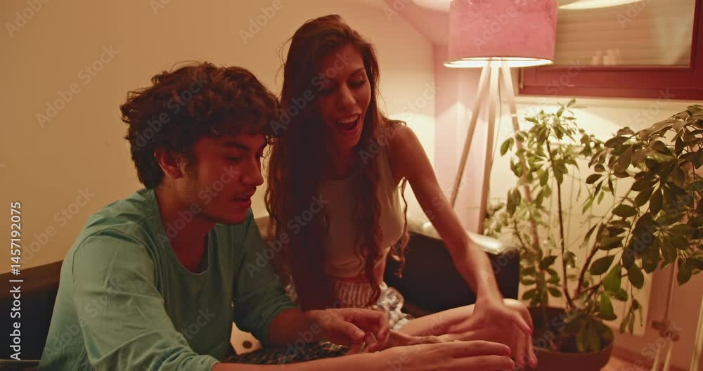 A young couple sits on their couch in their pajamas, playing Jenga and eating snacks. They smile and laugh, enjoying their time together.