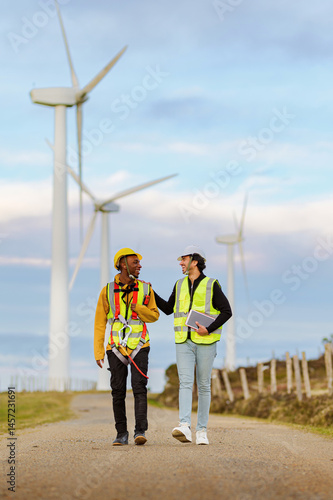 Obraz na płótnie Two engineers wearing safety vests and helmets walk along a path in a wind turbine park, discussing renewable energy projects and operations w salonie