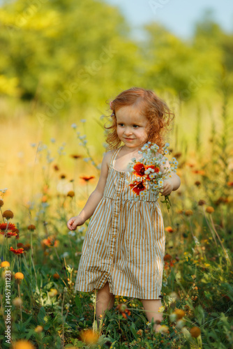 Little red-haired girl in a striped dress enjoying the scent of wildflowers on a sunny summer day. She is standing in a blooming field, holding a bouquet close to her nose and gently smelling it.