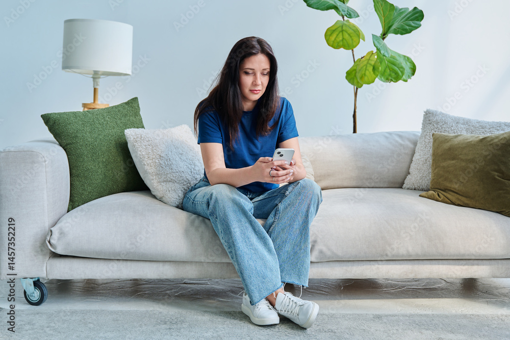 Serious young woman sitting at home on the couch using smartphone