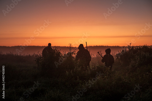 Group of hunters with shotguns going through rural field towards forest during sunset with orange sky and forest on horizon 