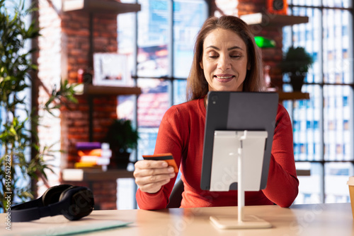 Caucasian adult at home pays for an order online on her tablet, entering credit card digits to confirm a transaction and make payment. Casual happy woman does online shopping in her loft.