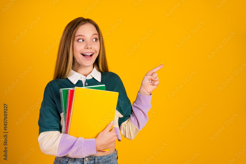 Happy young student holding colorful notebooks pointing in excitement against a vibrant yellow background