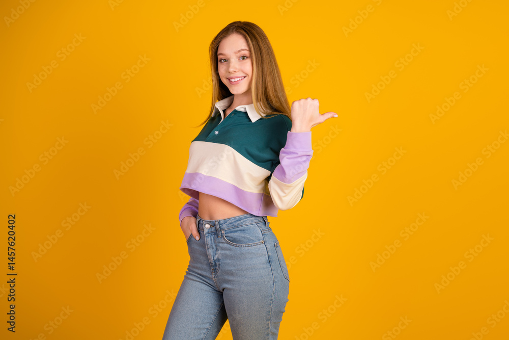 Young woman in casual colorful pullover pointing back with a cheerful smile, yellow backdrop.