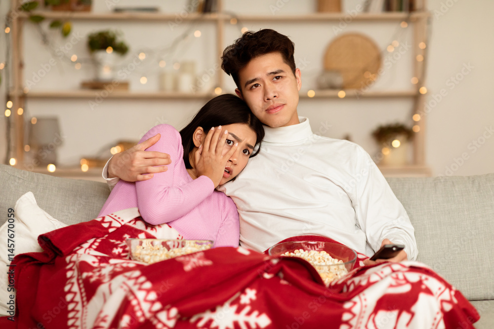 Obraz premium Japanese Couple Watching Scary Movie On TV Spending Christmas Eve Sitting On Sofa In Front Of Television Set In Living Room At Home, Covered With Blanket. Family Watches Horror During Winter Holidays