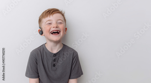Laughing young boy with light skin, red hair, and freckles wearing hearing aid. Dressed in grey t-shirt, smiling broadly with missing teeth. Light plain background with copy space on the right


