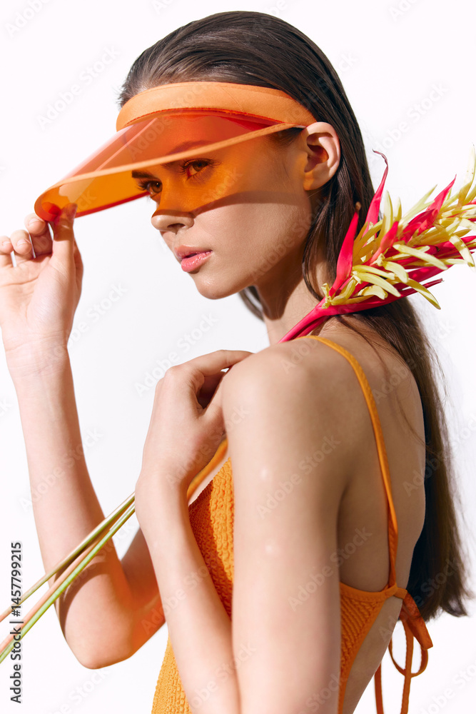Young woman wearing bright orange outfit with visor, holding tropical flowers in a minimalist studio setting, showcasing summer fashion and vibrant emotions.