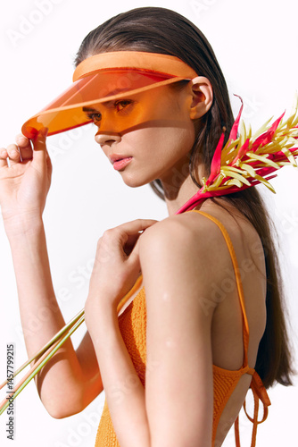 Obraz na płótnie Young woman wearing bright orange outfit with visor, holding tropical flowers in a minimalist studio setting, showcasing summer fashion and vibrant emotions. w salonie