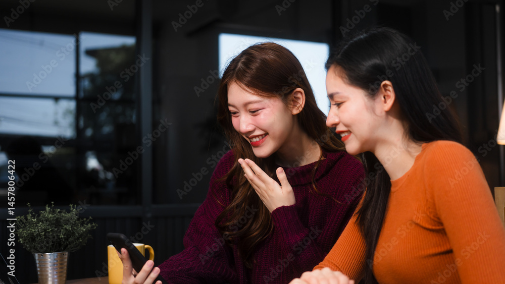 Two Asian women are sitting at a kitchen table, learning online at home. They smile happily while using laptops, surrounded by fruits and a coffee cup, enjoying their time