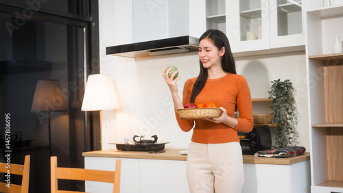 An Asian woman stands in the kitchen holding a basket of fresh fruits, ready to prepare a healthy meal. The kitchen is bright, and she radiates a sense of vitality
