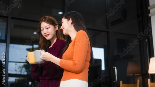Two Asian women are sitting at a kitchen table, learning online at home. They smile happily while using laptops, surrounded by fruits and a coffee cup, enjoying their time