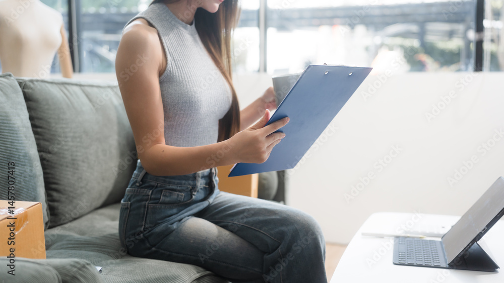 Happy Asian woman packing parcel boxes at a sofa table, smiling joyfully while checking online orders on laptop.She prepares products for delivery,managing her successful home-based online business