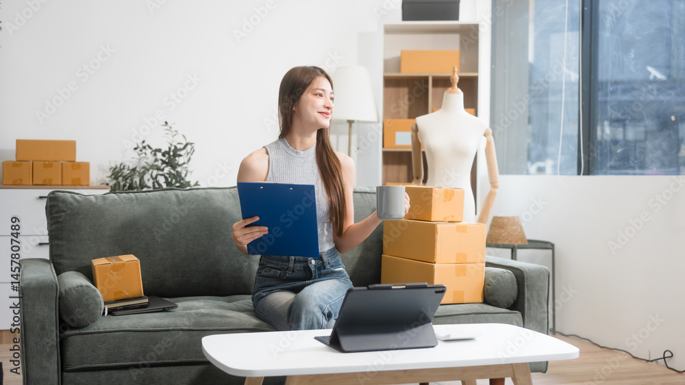 Happy Asian woman packing parcel boxes at a sofa table, smiling joyfully while checking online orders on laptop.She prepares products for delivery,managing her successful home-based online business Happy Asian woman packing parcel boxes at a sofa table, smiling joyfully while checking online orders on laptop.She prepares products for delivery,managing her successful home-based online business