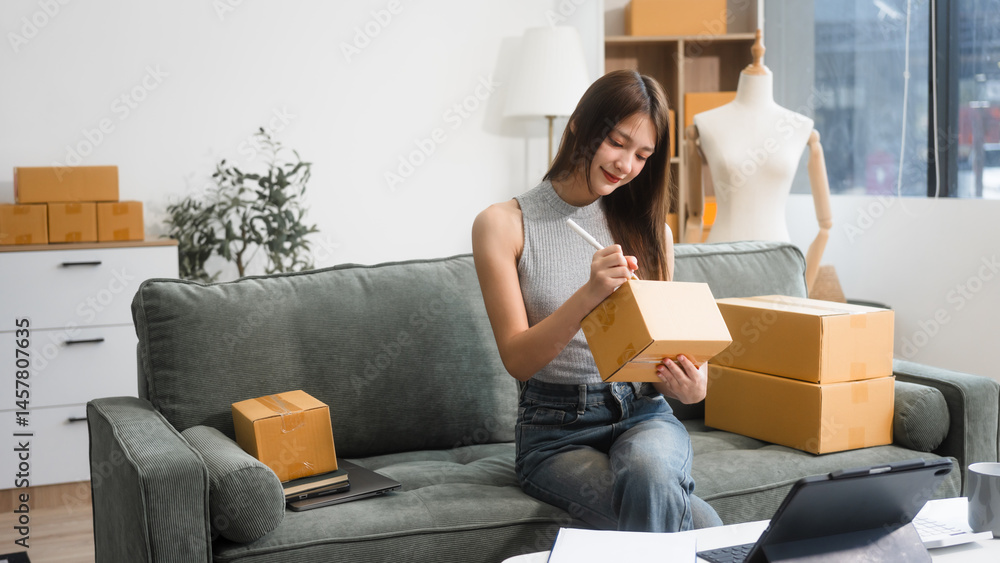 Happy Asian woman packing parcel boxes at a sofa table, smiling joyfully while checking online orders on laptop.She prepares products for delivery,managing her successful home-based online business