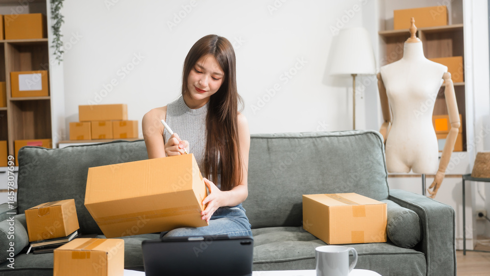 Happy Asian woman packing parcel boxes at a sofa table, smiling joyfully while checking online orders on laptop.She prepares products for delivery,managing her successful home-based online business