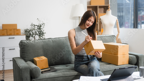 Obraz na płótnie Happy Asian woman packing parcel boxes at a sofa table, smiling joyfully while checking online orders on laptop.She prepares products for delivery,managing her successful home-based online business w salonie