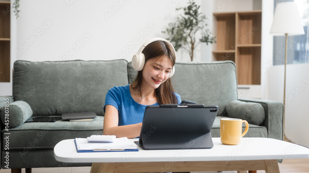 Asian woman sitting at a sofa table, wearing headphones and learning online at home.Focused on her laptop screen, she studies comfortably in a cozy environment,embracing digital education technology Asian woman sitting at a sofa table, wearing headphones and learning online at home.Focused on her laptop screen, she studies comfortably in a cozy environment,embracing digital education technology