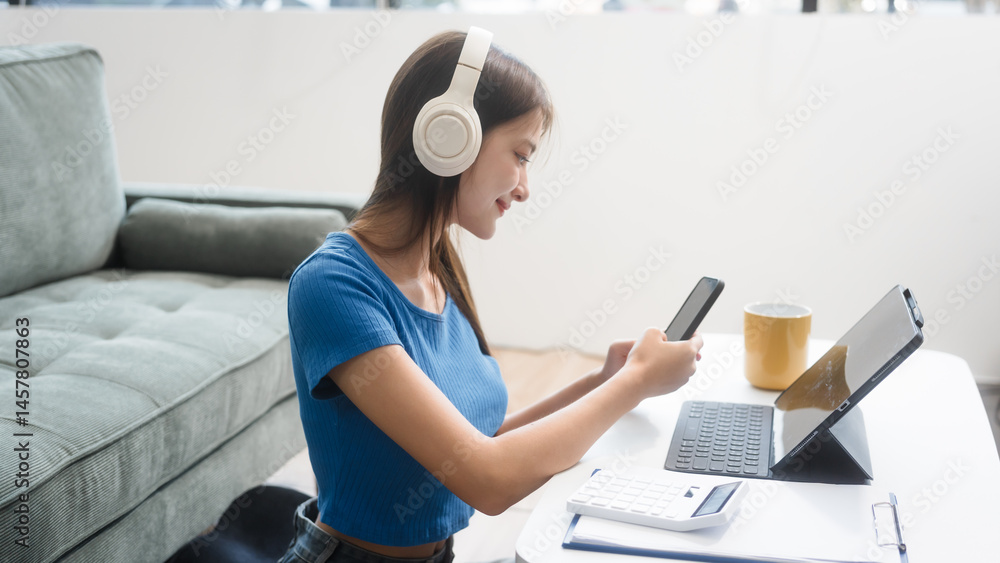 Asian woman sitting at a sofa table, wearing headphones and learning online at home.Focused on her laptop screen, she studies comfortably in a cozy environment,embracing digital education technology