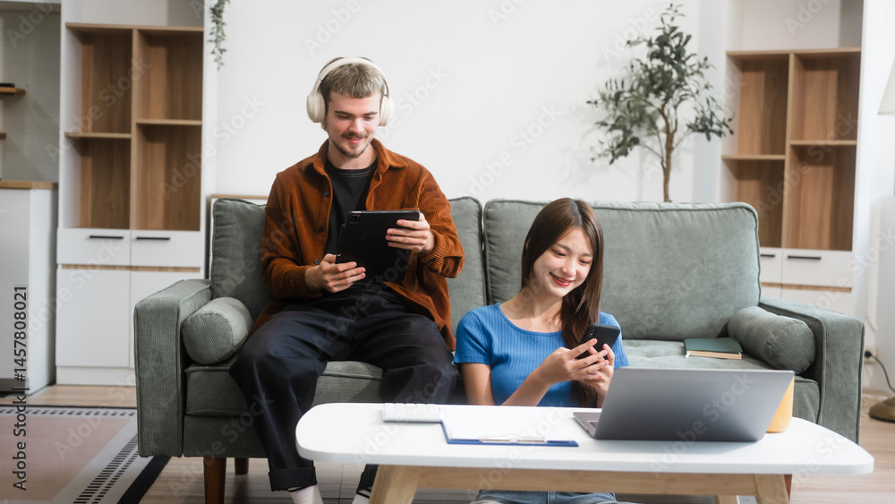 A Caucasian man and an Asian woman sit together at a table near the sofa, using a laptop at home. They appear relaxed and focused, working or browsing the internet together