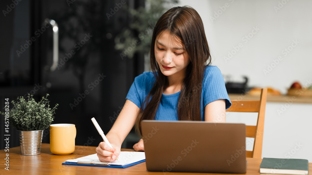 An Asian woman sits comfortably at her home desk, focused on her laptop. She works online in a relaxed setting, managing tasks efficiently with dedication, concentration, and a positive attitude