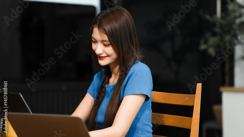 Obraz na płótnie An Asian woman sits comfortably at her home desk, focused on her laptop. She works online in a relaxed setting, managing tasks efficiently with dedication, concentration, and a positive attitude w salonie