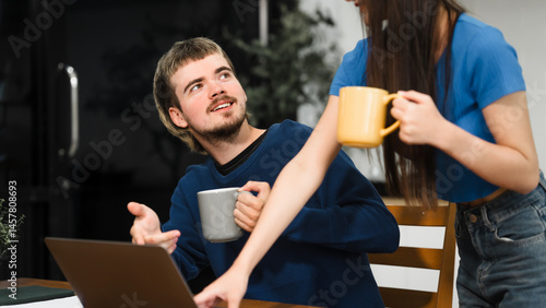 Caucasian man and an Asian woman sit at a table at home, engaged in online learning. They share ideas, talk casually, and sip coffee, creating a relaxed and focused atmosphere