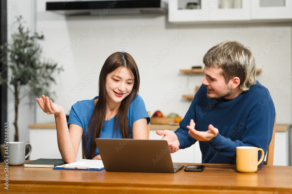 Caucasian man and an Asian woman sit at a table at home, engaged in online learning. They share ideas, talk casually, and sip coffee, creating a relaxed and focused atmosphere
