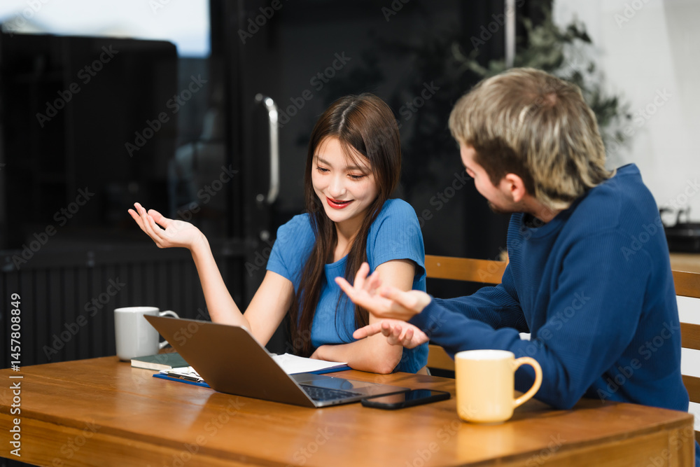 Caucasian man and an Asian woman sit at a table at home, engaged in online learning. They share ideas, talk casually, and sip coffee, creating a relaxed and focused atmosphere
