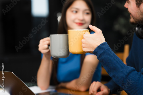 Obraz na płótnie Caucasian man and an Asian woman sit at a table at home, engaged in online learning. They share ideas, talk casually, and sip coffee, creating a relaxed and focused atmosphere w salonie